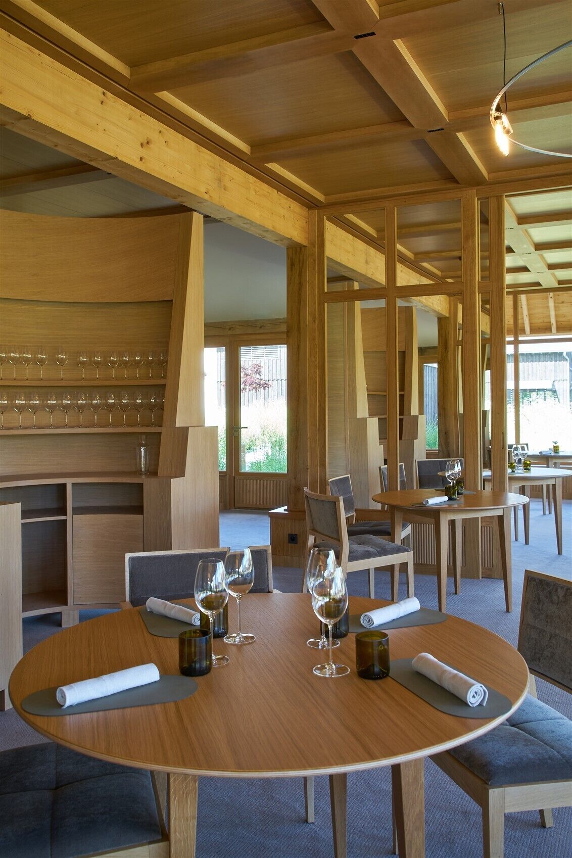 A modern dining area with wooden tables and chairs, glassware, and a view of greenery outside.