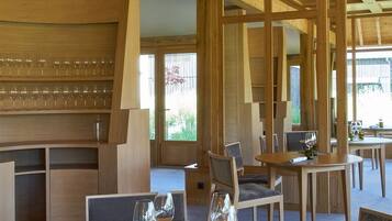 A modern dining area with wooden tables and chairs, glassware, and a view of greenery outside.