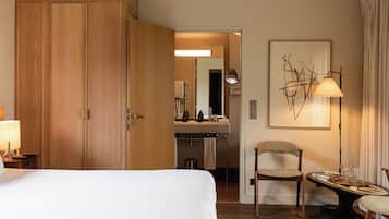 Quarto com uma cama grande, guarda-roupa de madeira, uma escrivaninha com cadeira, abajur e banheiro visível através de uma porta aberta.