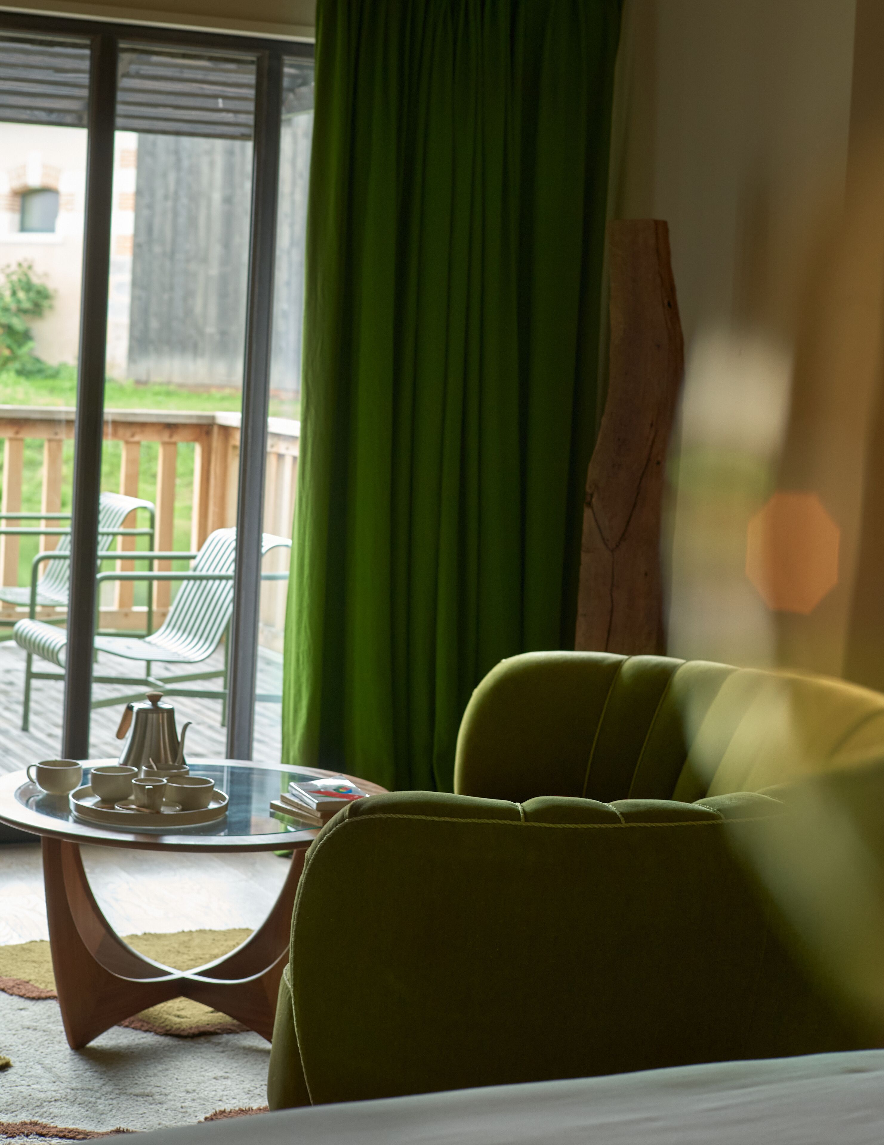 A room with a green armchair, a glass table with a tray of tea set, and a view of the outdoors through a window with green curtains.
