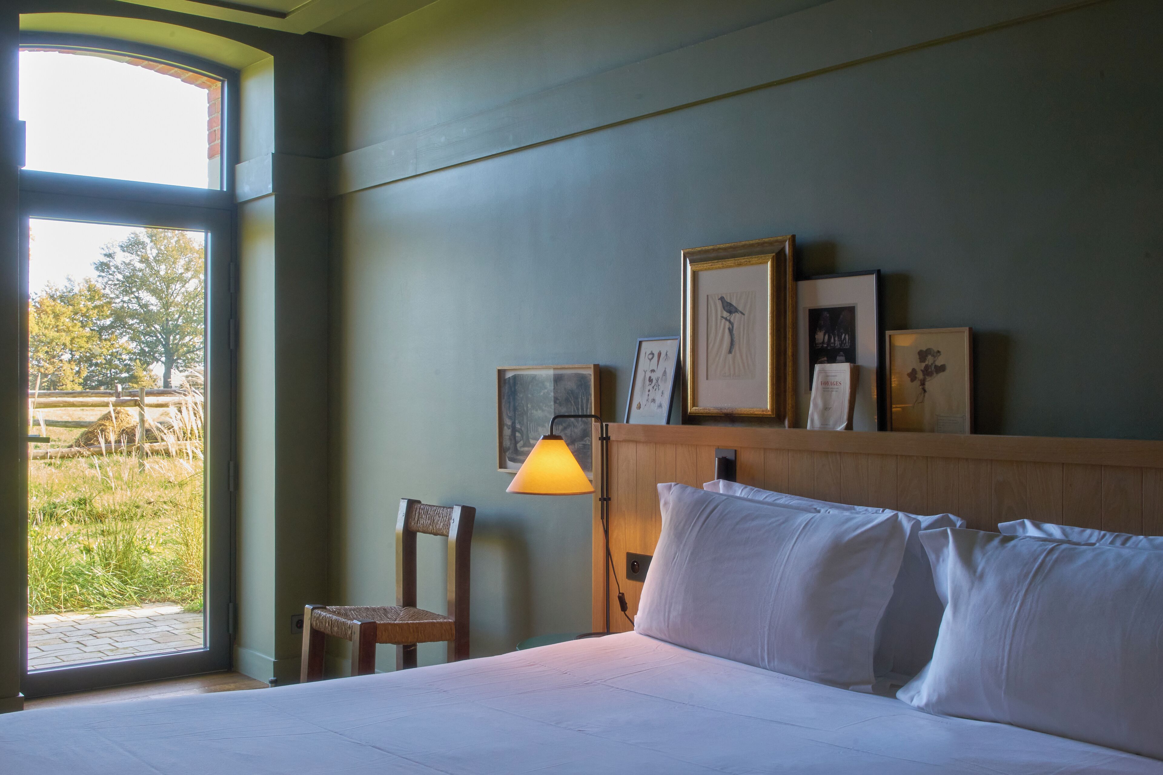A hotel room with a bed, a wooden headboard, a window with a view of trees, and a wall with framed pictures.