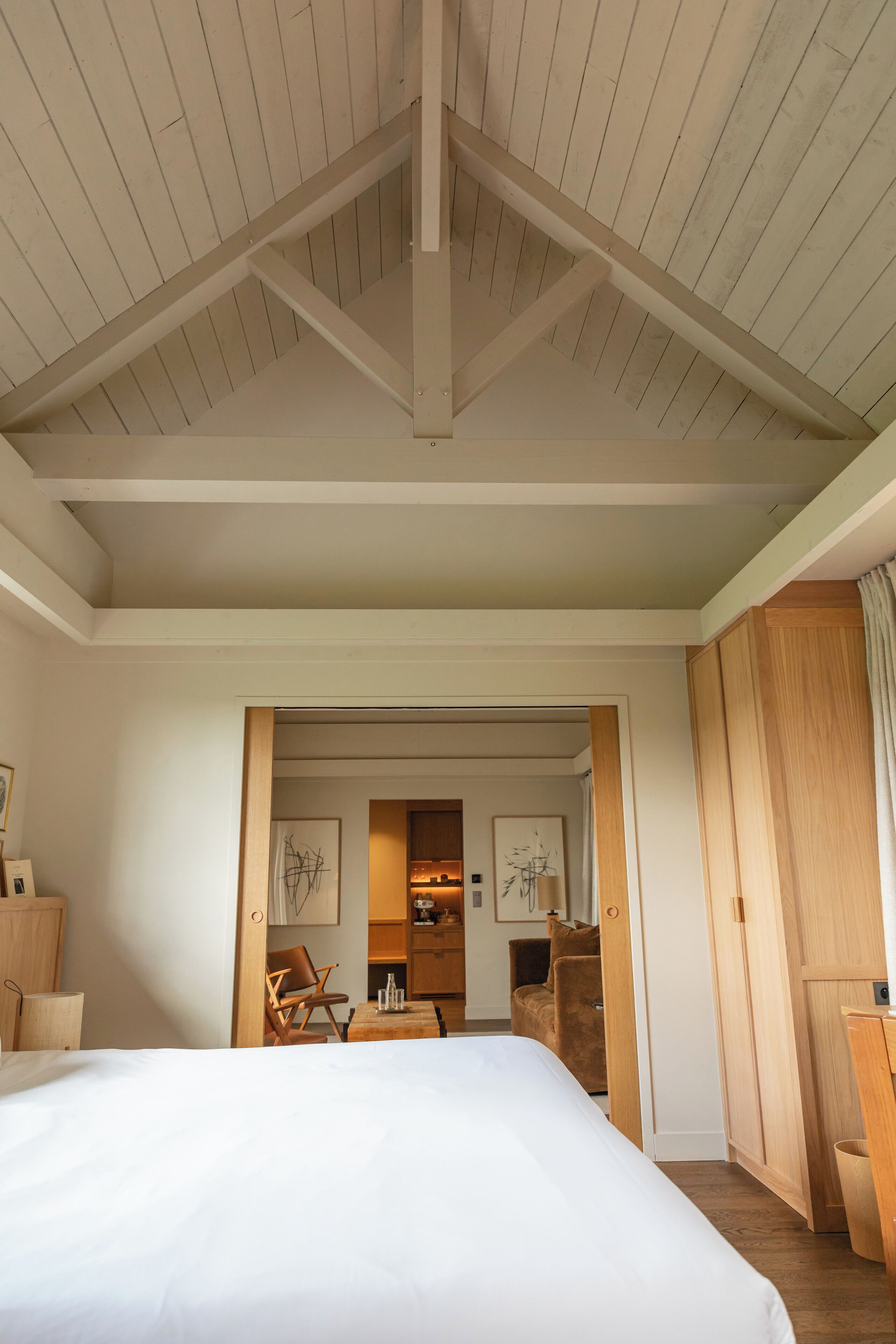 A bedroom with a large bed, wooden ceiling beams, a wooden wardrobe, and a desk with a chair.
