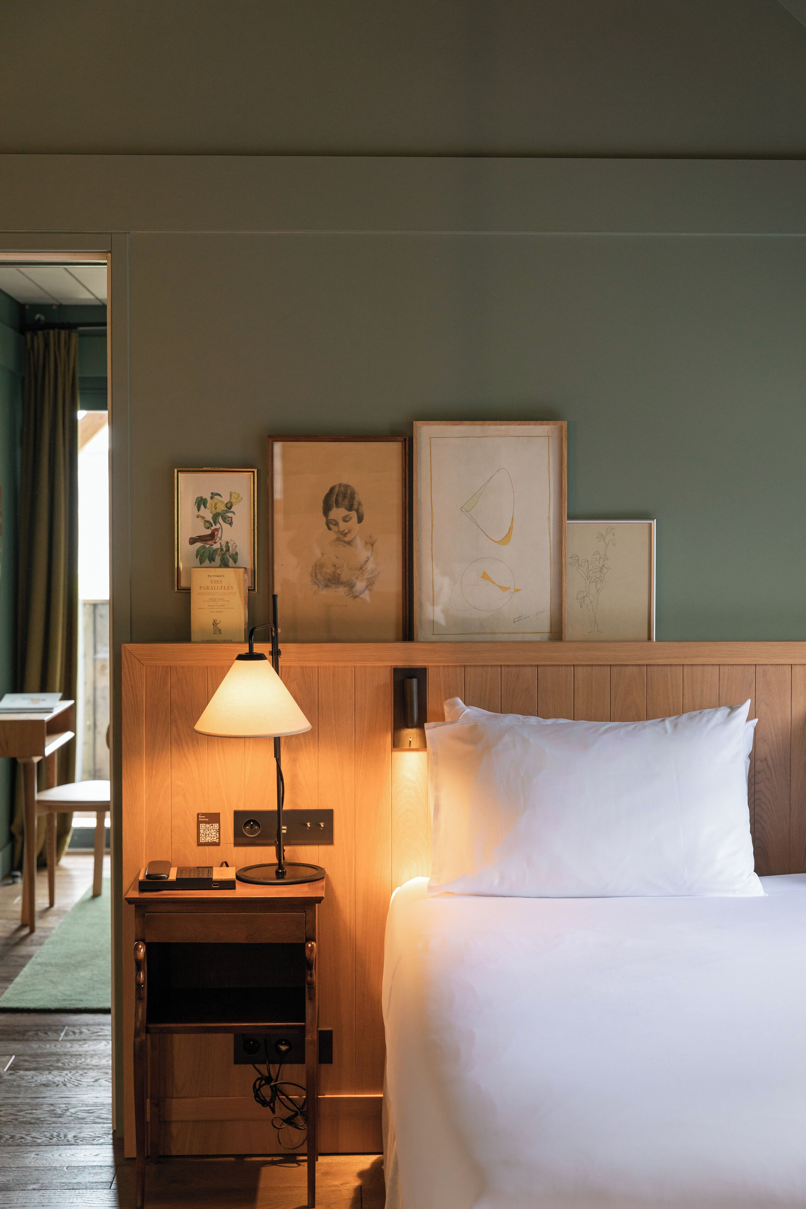Une chambre d’hôtel avec un lit, une table de chevet avec une lampe, une petite étagère avec des photos encadrées et une tête de lit en bois.
