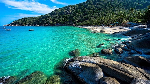 Ein Strand mit klarem türkisblauem Wasser, felsigem Ufer und einem bewaldeten Hügel im Hintergrund.