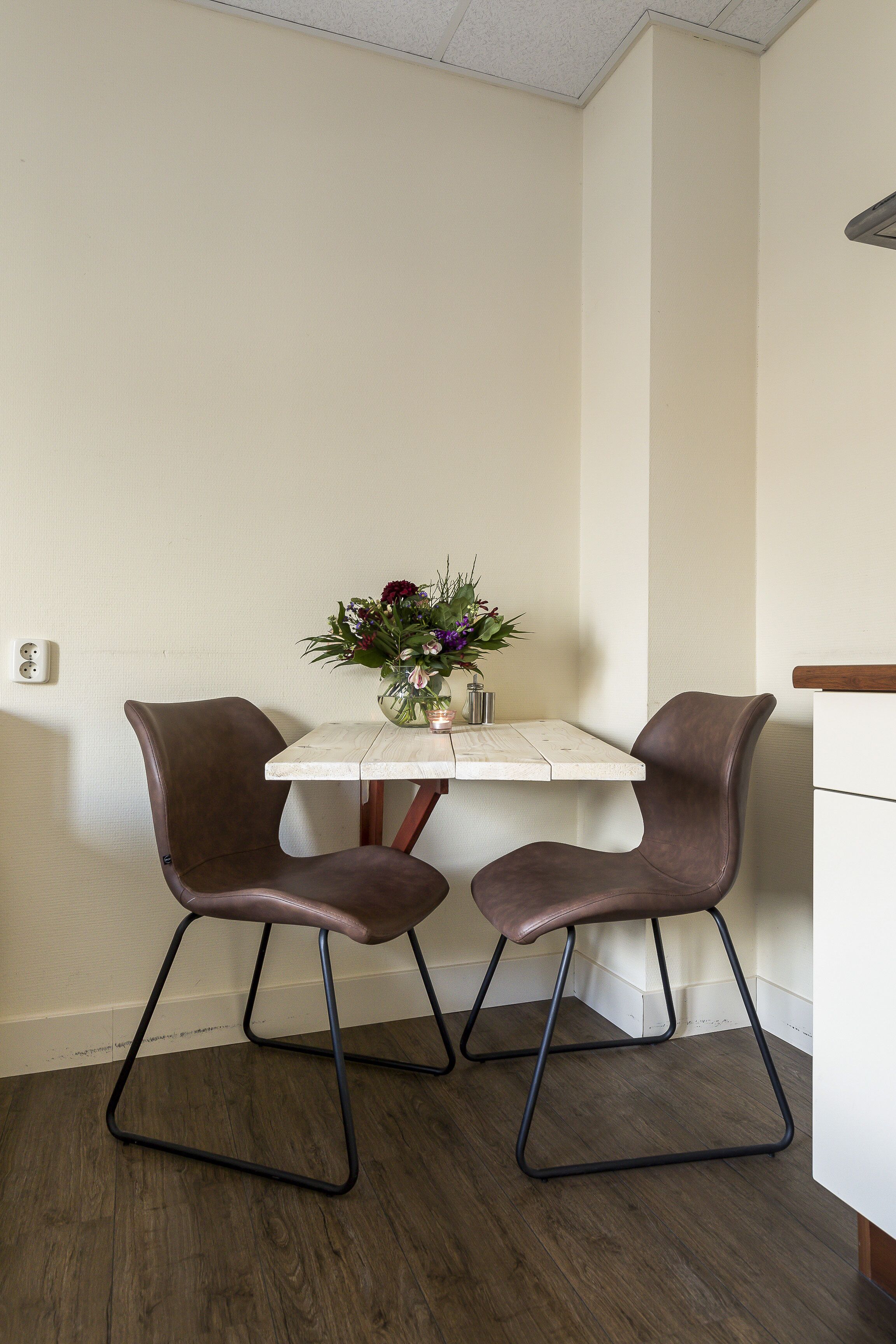 A corner room with a white table and two brown chairs. A vase with flowers is on the table.