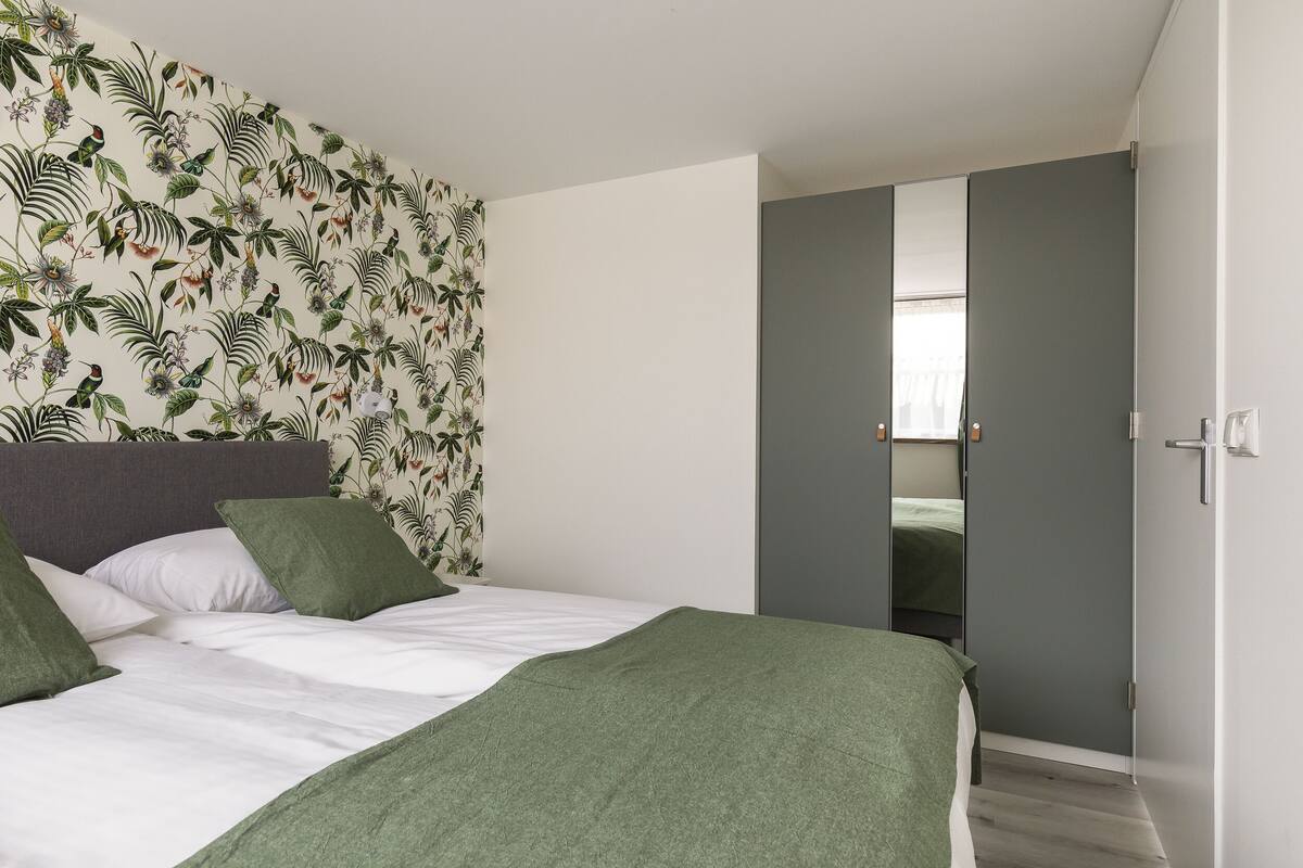 Un dormitorio con una cama, almohadas verdes, una manta verde, papel tapiz floral y un armario gris.