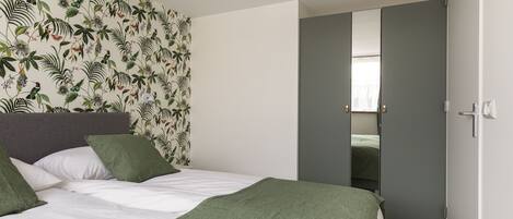 A bedroom with a bed, green pillows, a green blanket, a floral wallpaper, and a grey wardrobe.