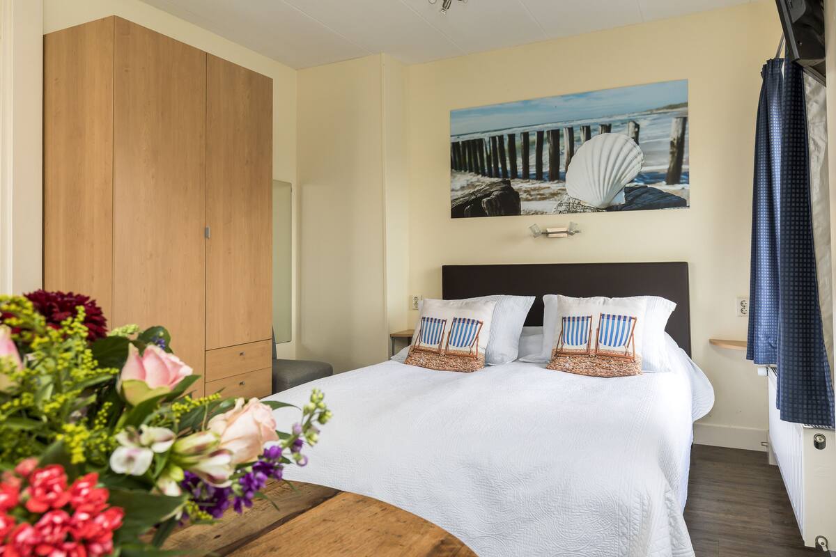 Un dormitorio con una cama, un armario de madera, un cuadro de una playa y un jarrón de flores.