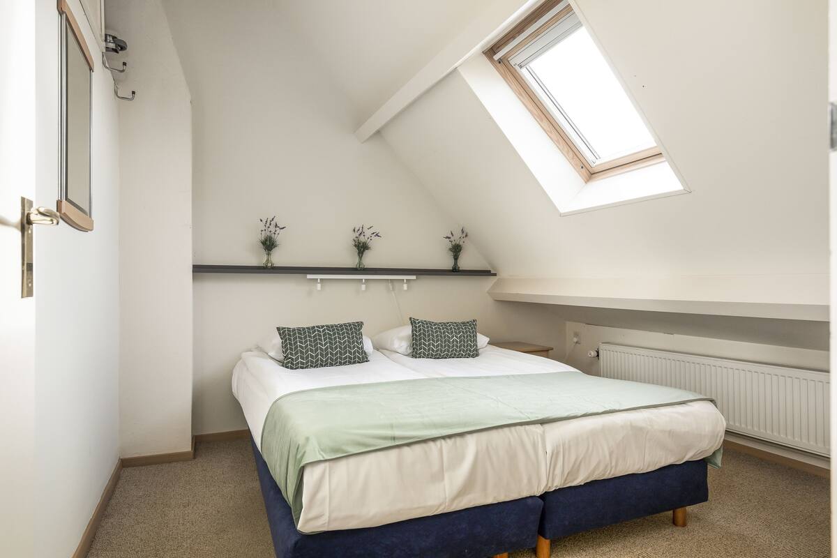 Un dormitorio con techo inclinado, tragaluz, cama con edredón verde y blanco, y estante con pequeñas plantas.