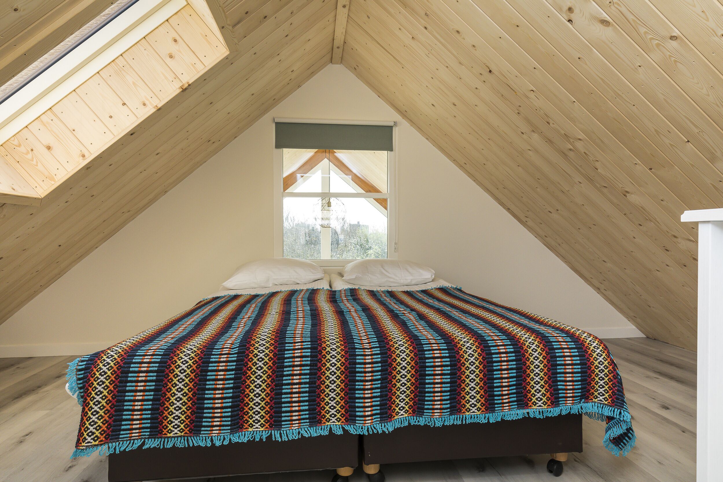 A bedroom with a sloped ceiling, a bed with a colorful blanket, two pillows, and a window with a view.
