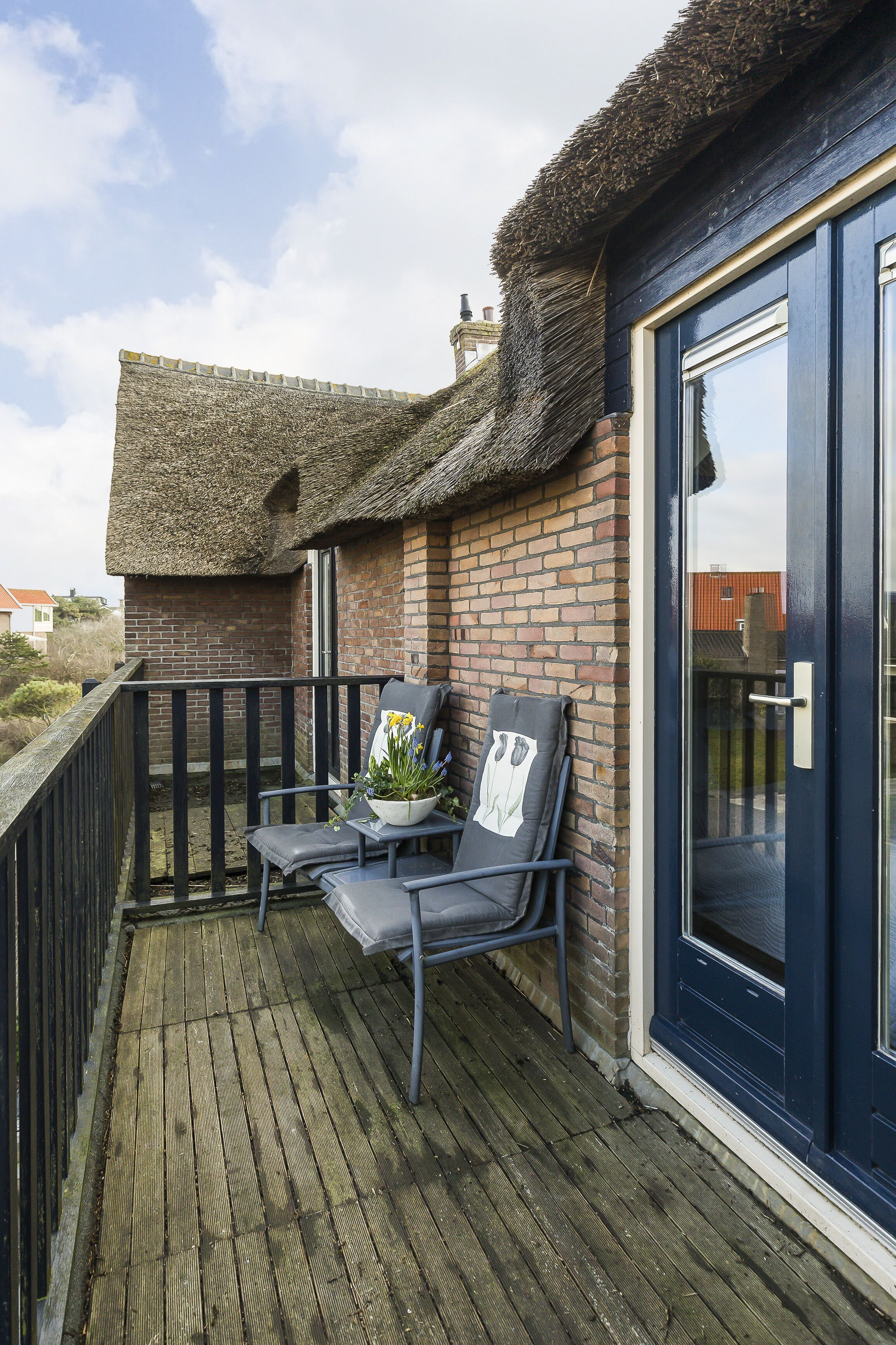 A balcony with a thatched roof, a brick wall, and a dark blue door. There are two chairs and a potted plant on the balcony.