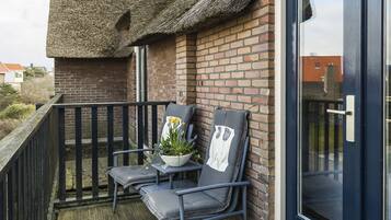 A balcony with a thatched roof, a brick wall, and a dark blue door. There are two chairs and a potted plant on the balcony.