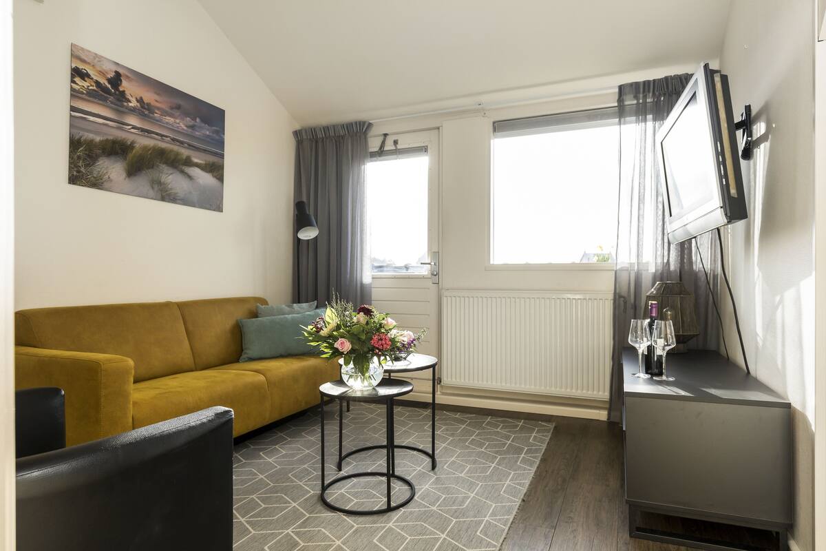 Una sala de estar con un sofá color mostaza, una pequeña mesa redonda con un jarrón de flores, un televisor sobre un mueble bajo y una ventana con cortinas.