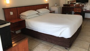 A hotel room with a large bed, wooden headboard, bedside tables, a desk with a chair, a television, and a mirror. - The Enclave (Francistown)