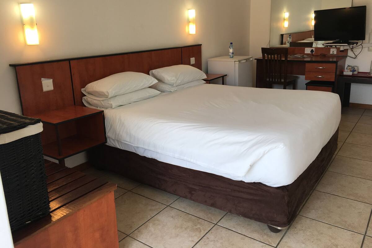 A hotel room with a large bed, wooden headboard, bedside tables, a desk with a chair, a television, and a mirror.