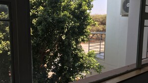 A view through a window with a metal railing, a tree, and a building with a white exterior. - The Enclave (Francistown)