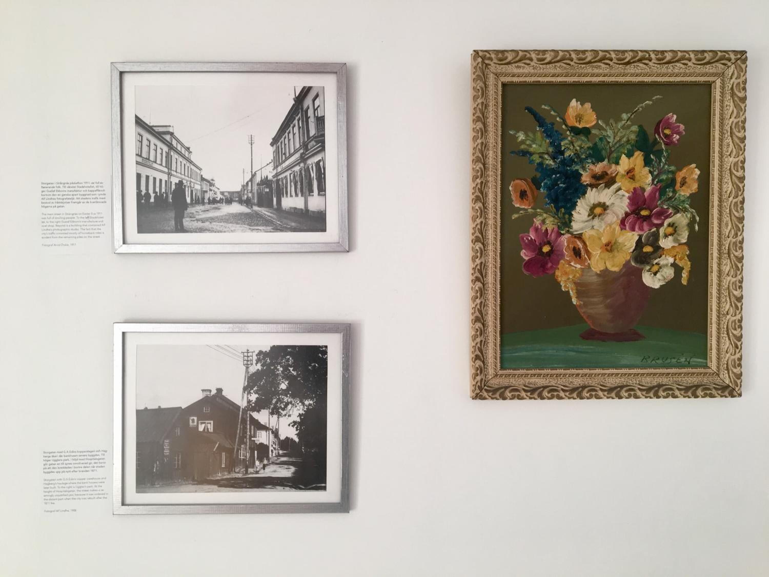 Drie ingekaderde foto’s aan een muur: een zwart-wit straatbeeld, een kleurrijke bloevas, en nog een zwart-wit straatbeeld.