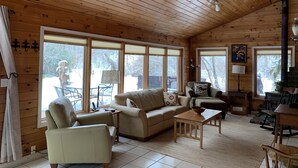 Una cabaña acogedora con paredes y techo de madera, una chimenea, un sofá, una mesa de centro y ventanas amplias con vistas al exterior.