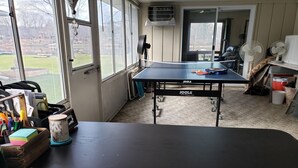 Ein Zimmer mit Tischtennisplatte, einem Sessel, einem Schreibtisch mit Computer und einem Fenster mit Blick auf einen Garten.
