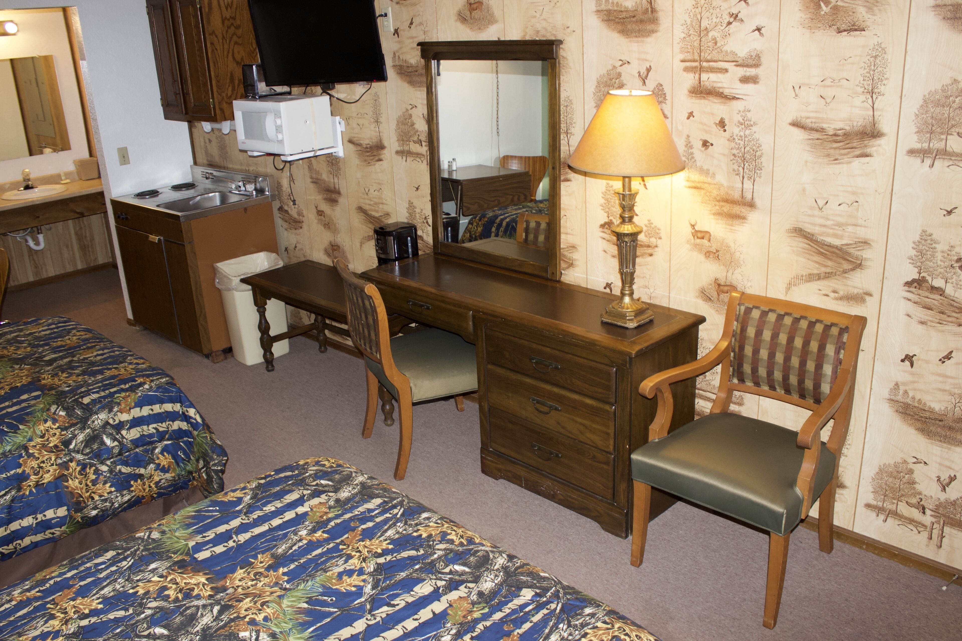 Une chambre d’hôtel avec un bureau en bois, une chaise, un lit avec une literie à motifs floraux, une lampe, un téléviseur et un miroir.