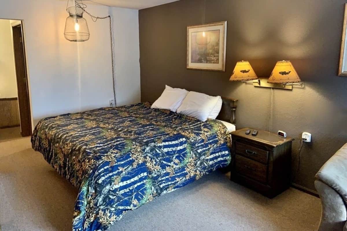 Un dormitorio con cama, dos lámparas de noche, una mesita de noche, un cuadro en la pared y una puerta a otra habitación.