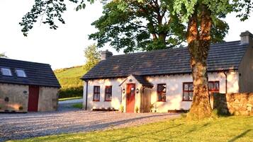 Une maison traditionnelle en pierre, avec une allée gravillonnée, une porte rouge et un grand arbre devant.