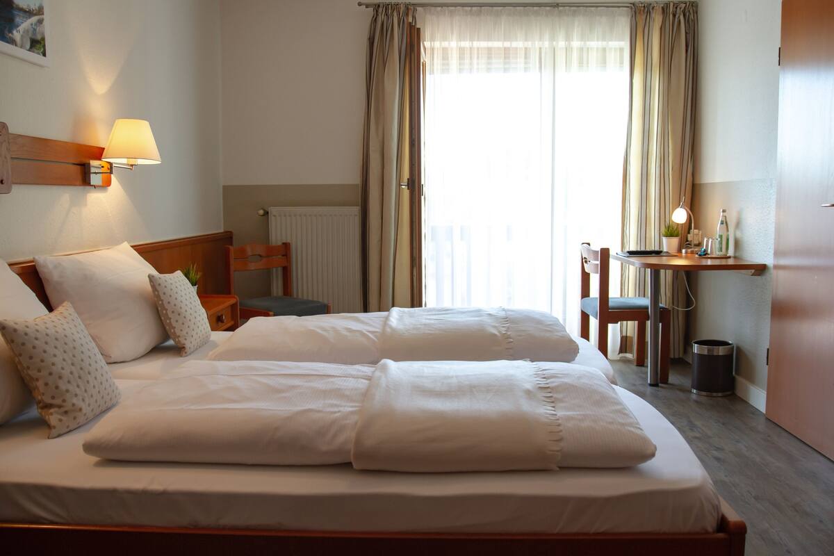 Un lit double avec une literie blanche, une table de chevet avec une lampe, une chaise, un petit bureau avec une lampe et un radiateur.