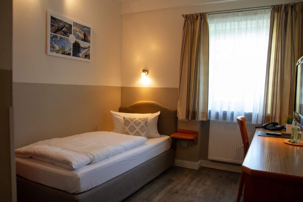 Une petite chambre d’hôtel avec un lit simple, un bureau avec une chaise, un radiateur et une fenêtre avec des rideaux.