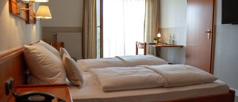 A hotel room with a double bed, bedside lamps, a desk with a lamp, a chair, and a TV mounted on the wall.