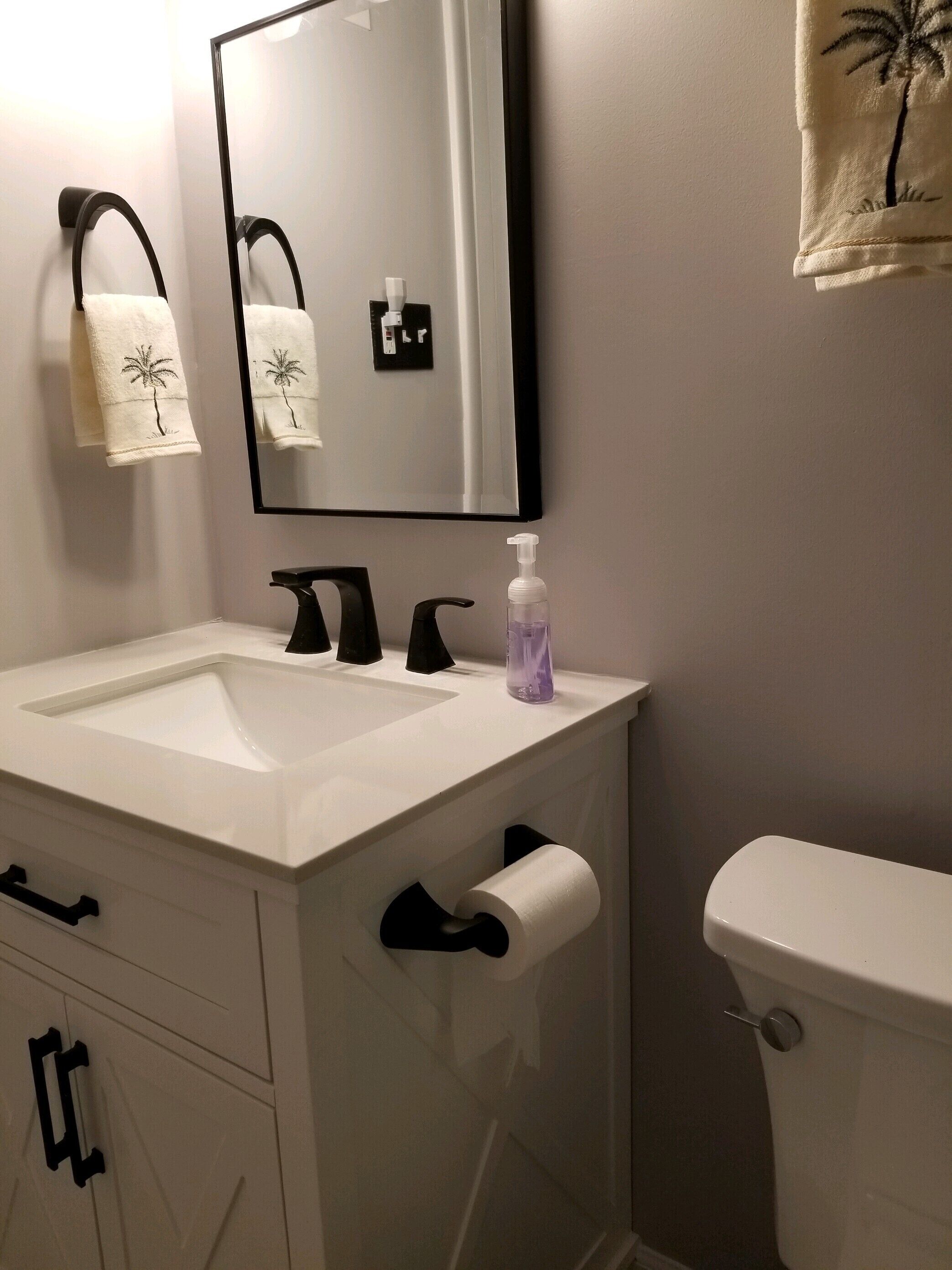 A bathroom with a white sink, black faucets, a mirror, a toilet, and a towel with a palm tree design.