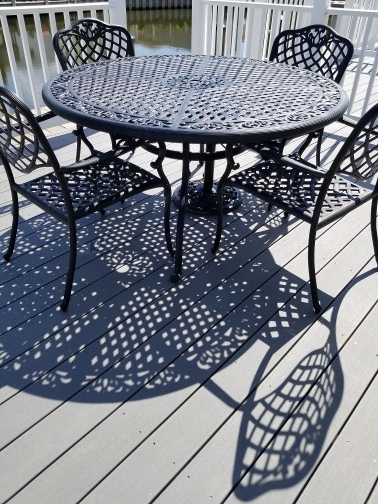 Una mesa metálica redonda con diseño de celosía, rodeada por cuatro sillas con un patrón similar, sobre una terraza de madera con barandilla blanca.