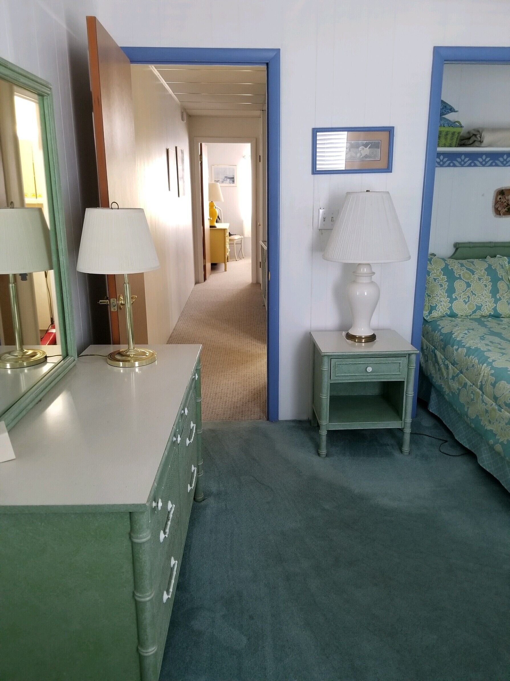 A hotel room with a green dresser, a bedside table with a lamp, a bed with floral bedding, and a hallway leading to another room.