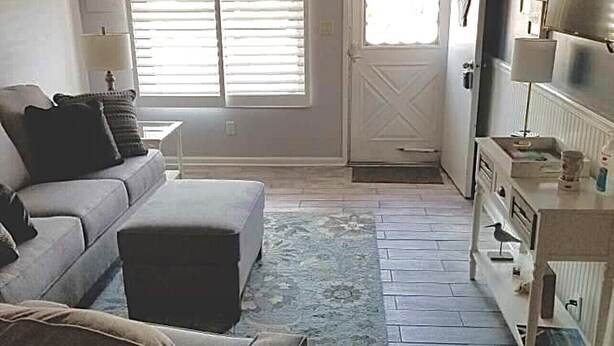 Ein Wohnzimmer mit Sofa, Hocker und Teppich. Eine Tür führt in einen anderen Raum, ein Fenster mit Jalousien ist ebenfalls zu sehen.
