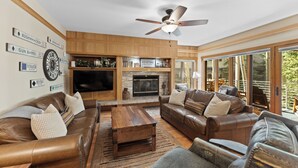 A spacious living room with leather sofas, a wooden coffee table, and a flat-screen TV. A fireplace is built into the wall, and there are large windows offering a view of the outdoors.