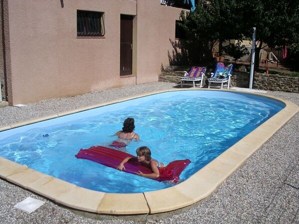 A swimming pool with two people in the water, surrounded by a pebble ground and a building with a window and door.