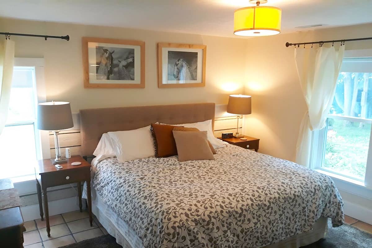 A bedroom with a bed, two bedside tables, a nightstand, a window with curtains, and two framed pictures on the wall.