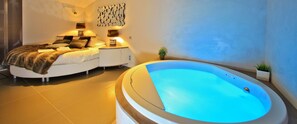Une chambre à coucher moderne avec un grand lit, des lampes de chevet, une œuvre d’art abstraite accrochée au mur, une petite plante en pot et une grande baignoire dotée d’un éclairage bleu.