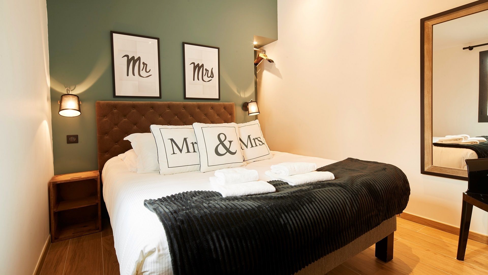 En snyggt bäddad säng med kuddar märkta med "Herr" och "Fru" i ett hotellrum med en sänggavel i trä, en spegel och inramade bilder på väggen.