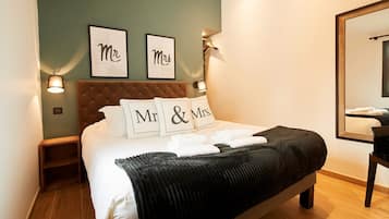 En snyggt bäddad säng med kuddar märkta med "Herr" och "Fru" i ett hotellrum med en sänggavel i trä, en spegel och inramade bilder på väggen.