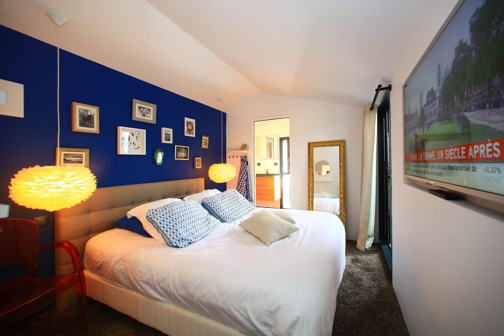Ett hotellrum med en stor säng, en röd fåtölj, en väggmonterad TV och inramade tavlor på den blå väggen.