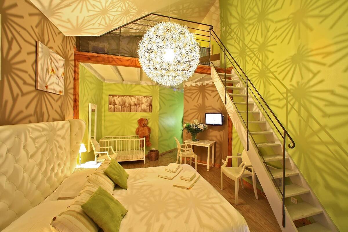 Ein Schlafzimmer mit einem großen Bett, einem Reisebettchen, einem Teddybären, einem Schreibtisch mit Computer und einer Treppe.