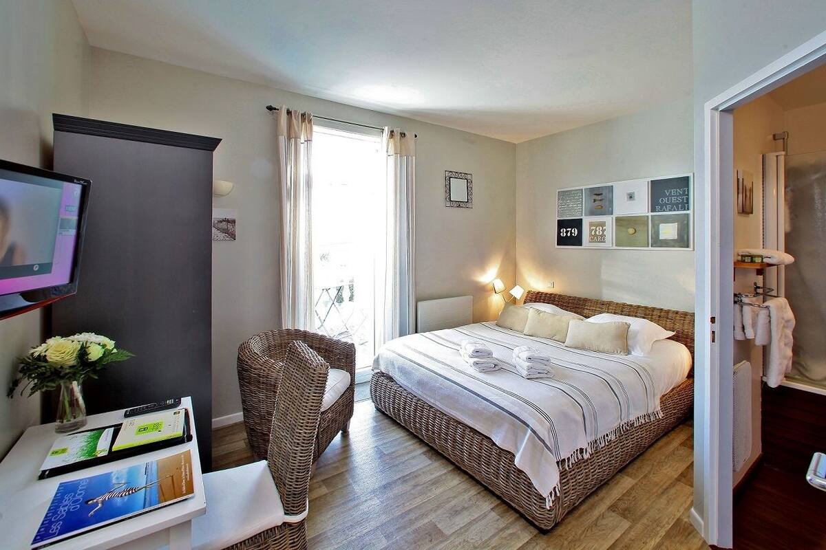 Ein Hotelzimmer mit einem Bett, einem Fernseher, einem Korbstuhl, einem kleinen Tisch mit Broschüren und einem Fenster mit Vorhängen.
