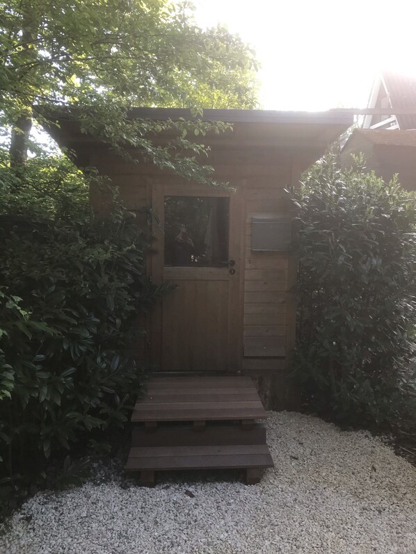 Une porte en bois avec une petite fenêtre, un escalier en bois menant à cette porte, et un chemin de gravier.