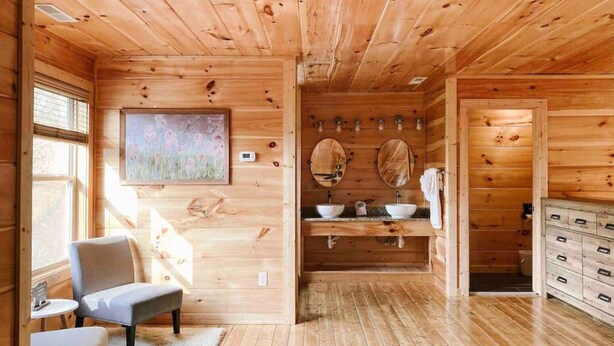 Ein Holzhaus-Interieur mit Holzboden, einer Bank, einem Gemälde, einem Waschbecken mit zwei Spiegeln und einem Schrank.