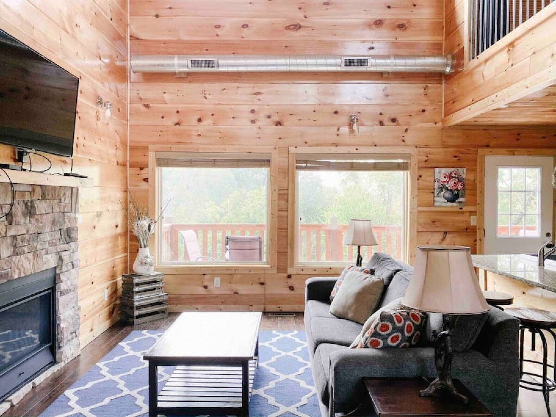 Ein gemütliches Blockhaus-Interieur mit Kamin aus Stein, Holzwänden, einer Couch mit gemusterten Kissen und einem Essbereich mit Lampe.