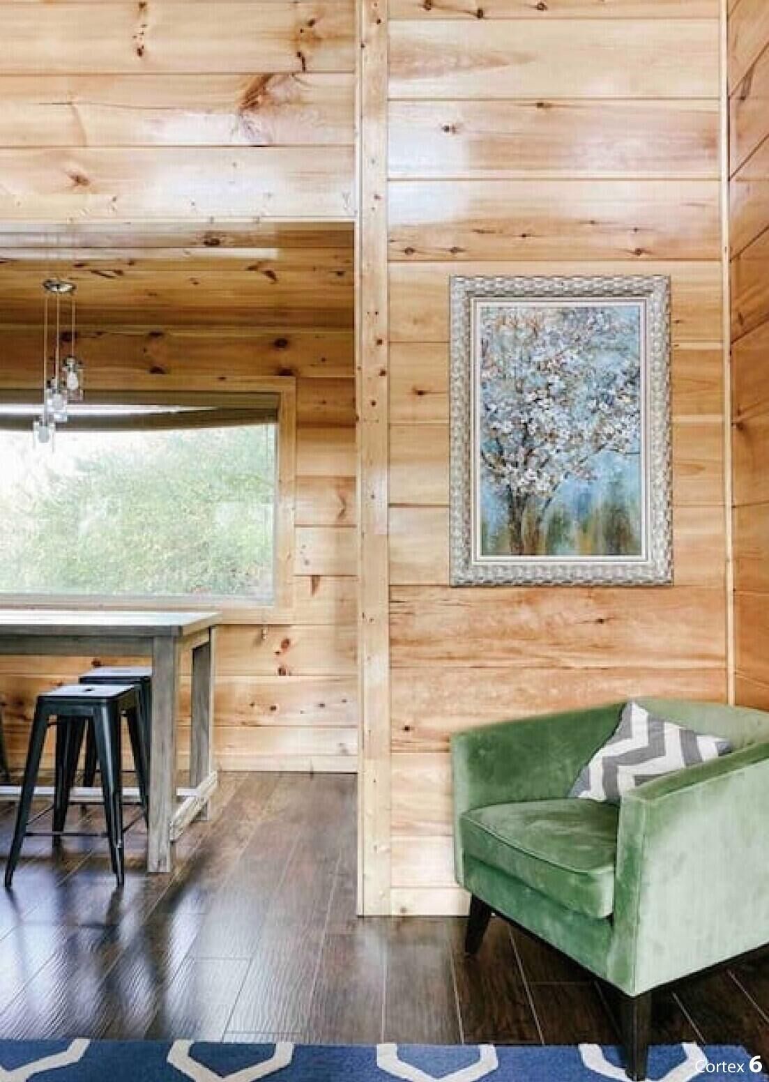 A wooden interior with a green armchair, a framed painting, and a dining area with stools.