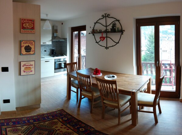 A dining area with a wooden table and chairs, a kitchen in the background, and a balcony with a view.