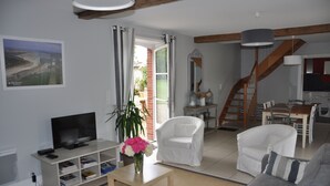A living room with a TV, white chairs, a sofa, a dining table, and a staircase.