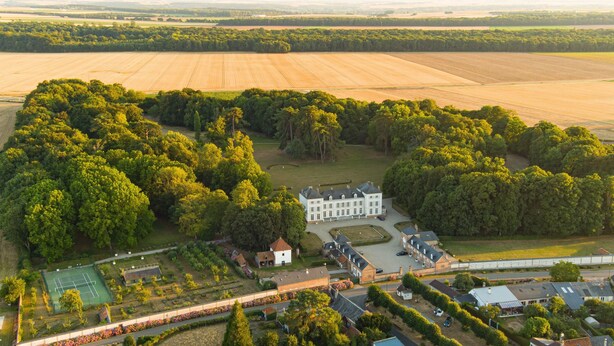 An aerial view of a large estate with expansive fields, dense forests, and a central white building with multiple wings.
