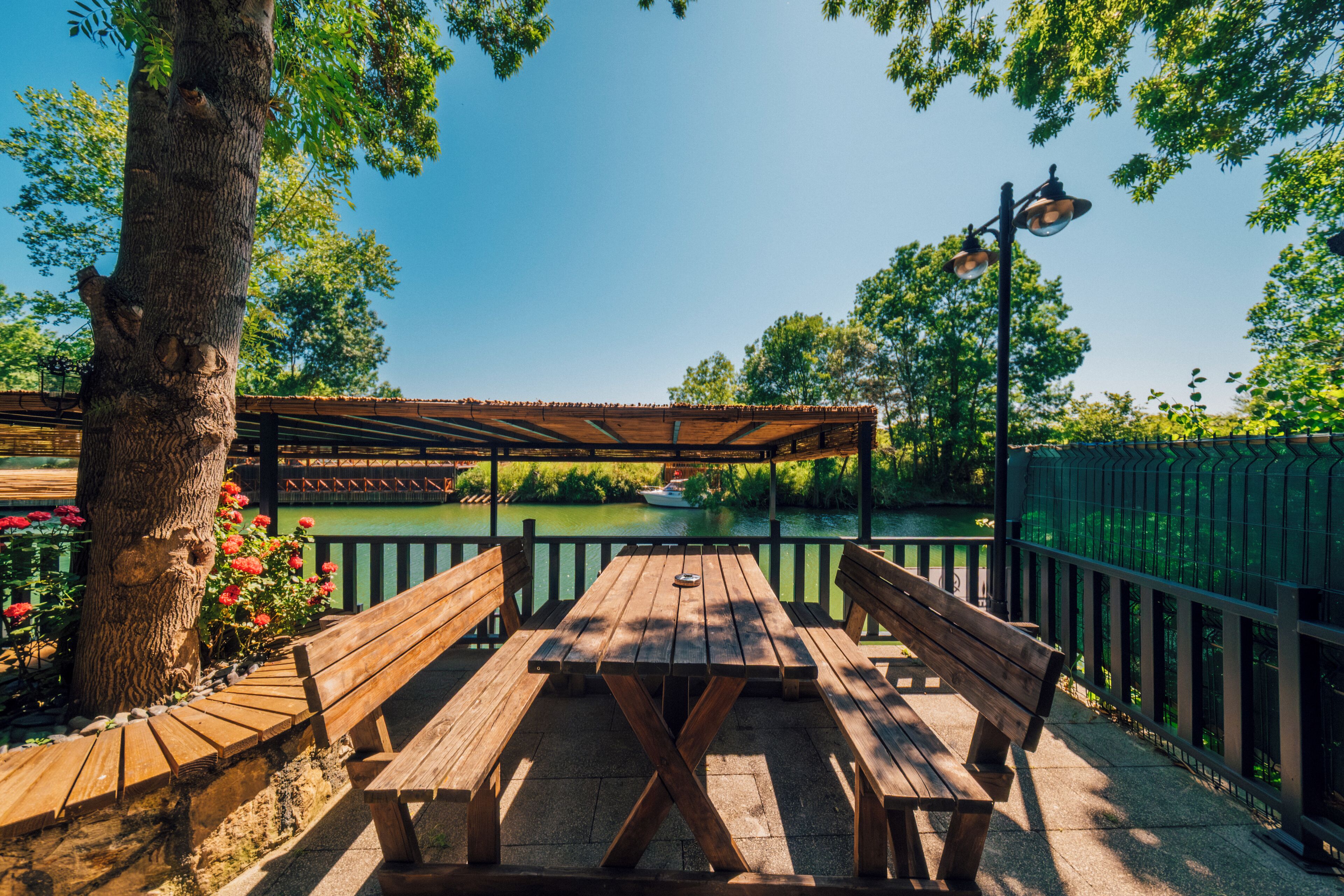 Een houten picknicktafel met banken, een pergola en uitzicht op een rivier met een boot.
